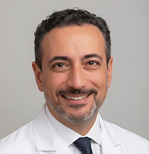 Mann mit lockigem Haar und Bart, in rosa Arztkleidung, lächelt in die Kamera.